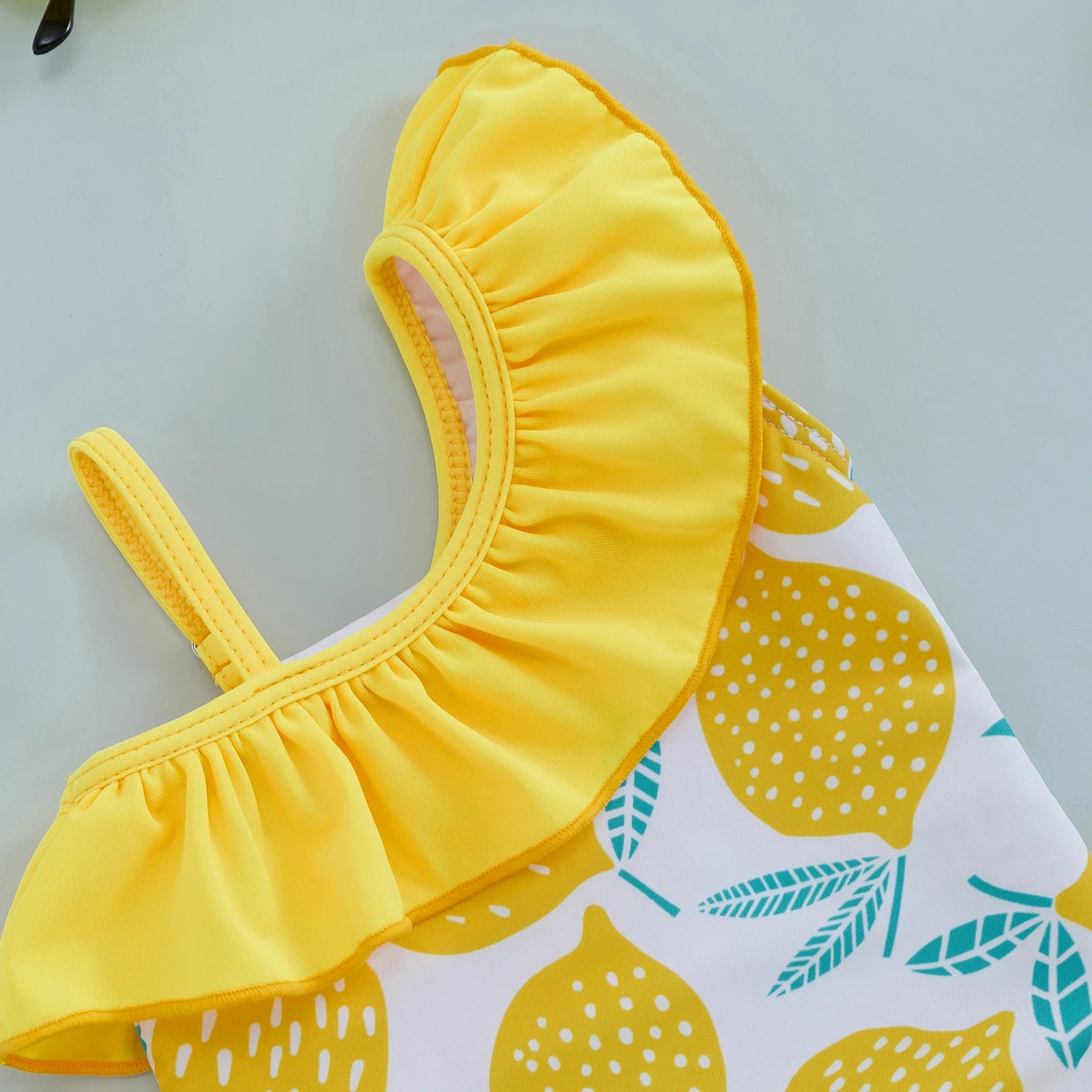 Baby girls one-shoulder swimsuit with a lemon print by Baby Bubble, featuring bright, colorful lemon-themed design and shoulder strap detail.